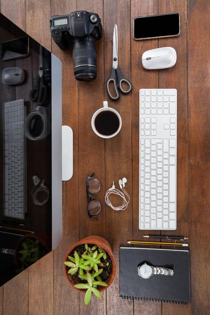 Overhead view of a neatly organized office desk featuring a computer, camera, smartphone, coffee cup, keyboard, mouse, scissors, glasses, earphones, plant, notebook, and watch. Ideal for illustrating productivity, modern workspace, office setup, and technology use in professional environments.