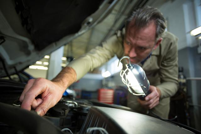 Mechanic examining car engine with lamp in repair garage. Ideal for illustrating automotive services, vehicle inspections, car maintenance, mechanic workshops, and professional repair services.