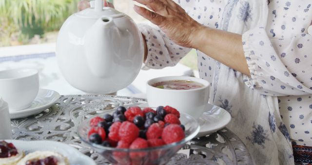 A senior Caucasian woman is pouring tea into a cup during a delightful afternoon tea, with copy space. Fresh berries and scones complement the elegant setting, evoking a sense of tradition and relaxation.