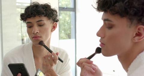 Man Applying Makeup with Brush While Checking Smartphone in Mirror