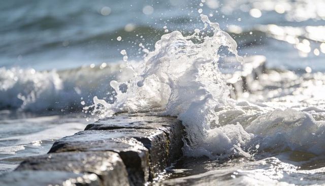 Crashing wave hitting stone groyne with sunlit spray and foamy surf on shoreline