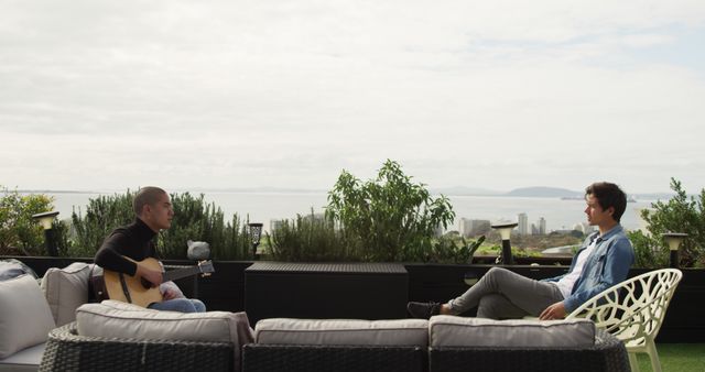 Two men are sitting on a patio with a stunning ocean view in the background. One of the men is playing a guitar while the other listens, creating a serene and leisurely atmosphere. Ideal for content related to leisure activities, outdoor relaxation, musical bonding, and scenic settings.