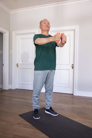 Senior chinese man doing gentle standing dumbbell workout on home mat in bright room