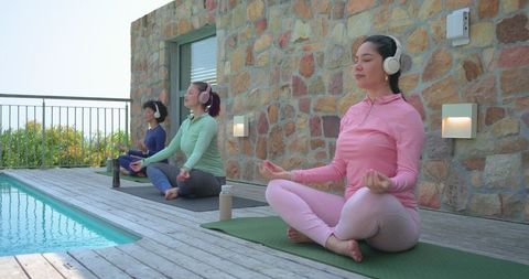 Diverse women meditating on pool deck wearing wireless headphones and pastel leggings