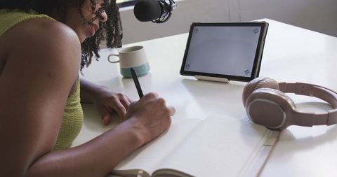 African american woman writing notes while recording podcast at home workspace tablet mic