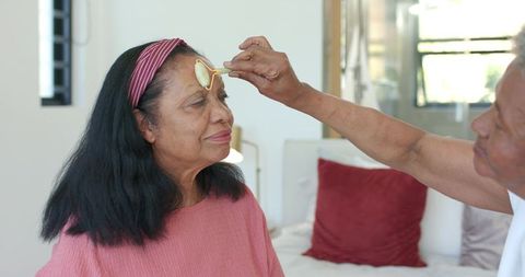 Senior woman receiving facial roller massage from caregiver in cozy bedroom wellness scene