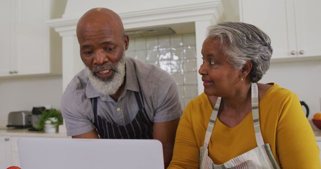 African american senior couple wearing aprons having a image call on laptop in the kitchen at home. retirement senior couple lifestyle living concept