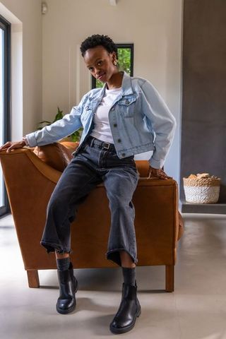 African american woman posing on leather armchair wearing denim jacket in minimalist living room