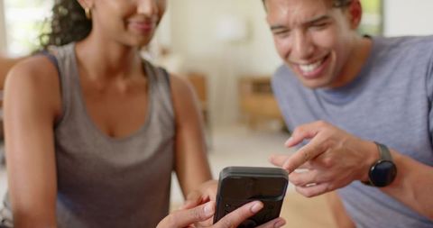 African couple laughing and sharing smartphone while pointing at screen in living room