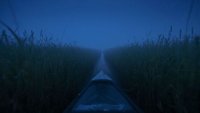 Kayak bow gliding through foggy marsh channel predawn calm reflective water and reeds