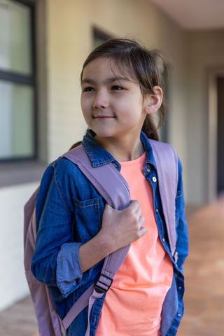 Asian elementary student holding backpack strap wearing denim jacket and coral tee