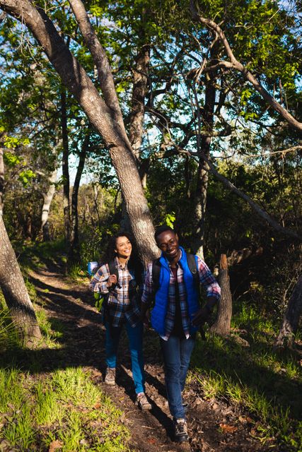 African American couple walking through forest while trekking, carrying backpacks and smiling. Ideal for use in travel blogs, adventure magazines, outdoor activity promotions, and healthy lifestyle campaigns.