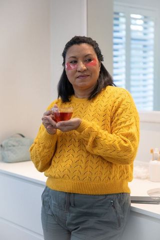 Mature woman enjoying morning self-care with pink under-eye patches and glass cup of tea