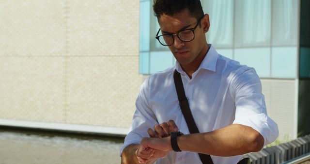Businessman wearing a white shirt and glasses checking the time on his smartwatch outdoors. Suitable for concepts such as punctuality, time management, business lifestyle, technology in professional life, and work-life balance.