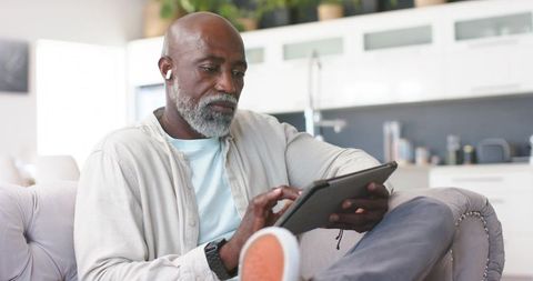 Senior african man relaxing on sofa using tablet with wireless earbuds home tech lifestyle