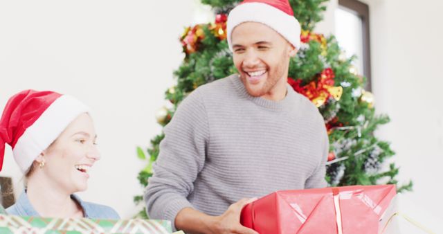 Capture of couple celebrating Christmas by exchanging gifts in front of decorated tree. Both wearing Santa hats and smiling, capturing holiday joy and warmth. Perfect for holiday marketing campaigns, social media posts, festive greeting cards, and promotional materials highlighting togetherness and joy during the holiday season.