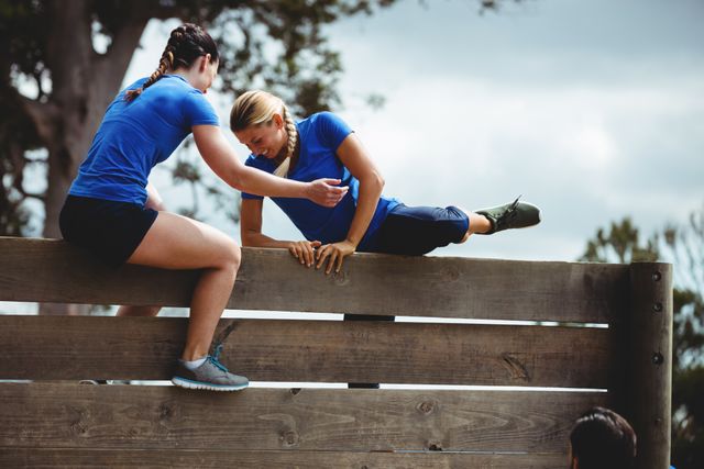 Female trainer assisting woman to climb a wooden wall during obstacle course