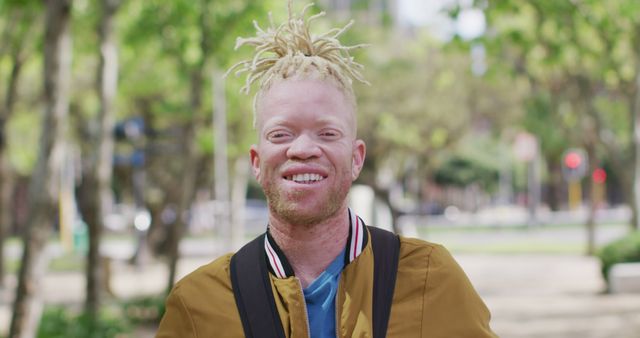 Young man with blond dreadlocks walking through park on a sunny day, relaxed and cheerful expression. Ideal for urban lifestyle, outdoor activities, positivity themes, and modern casual fashion adverts.
