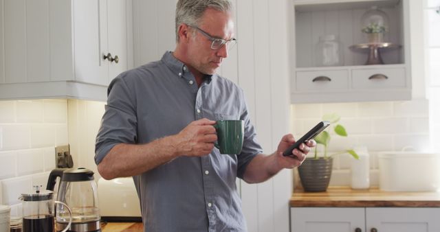Happy caucasian man using smartphone and drinking coffee in kitchen. Spending time at home alone.