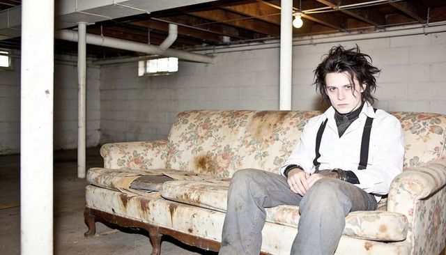 Brooding young man wearing white shirt and black suspenders sitting on worn floral sofa in basement