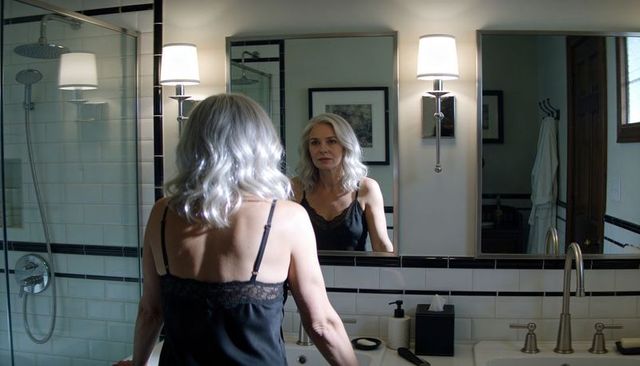 Mature woman standing at bathroom vanity looking in mirror during quiet self-care routine