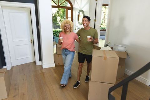 Diverse couple walking arm-in-arm carrying coffee amid unpacking boxes in new home foyer