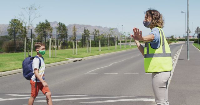 Visual showing a school crossing guard assisting a young student at a pedestrian crossing. Guard wears a safety vest and mask, child also wearing mask, suggesting focus on safety and social distancing. Ideal for content relating to child safety, educational institutions, community assistance, or traffic safety campaigns.