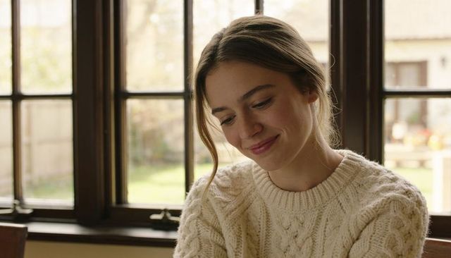 Woman sitting by sunlit paned window wearing cream cable-knit sweater, cozy portrait