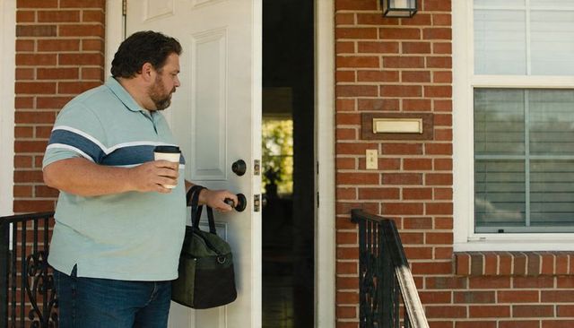 Man carrying duffel and coffee while opening front door on brick porch morning routine