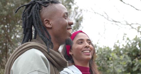 Diverse couple smiling and looking right while standing in park, candid outdoor portrait