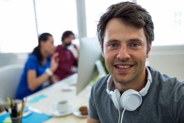 Young male graphic designer smiling at camera in a modern office environment. He is wearing headphones around his neck, indicating a creative and relaxed atmosphere. In the background, two colleagues are collaborating at a computer. This image is ideal for use in articles or advertisements related to creative professions, modern workspaces, teamwork, and professional environments.