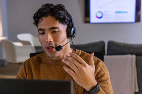 Young man in 20s wearing headset speaking and gesturing on laptop for remote meeting