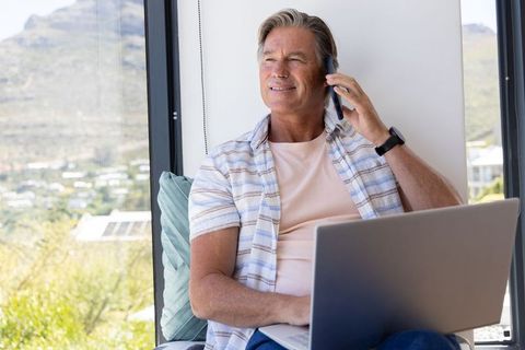 Mature man working remotely by window with laptop and phone, relaxed modern home office