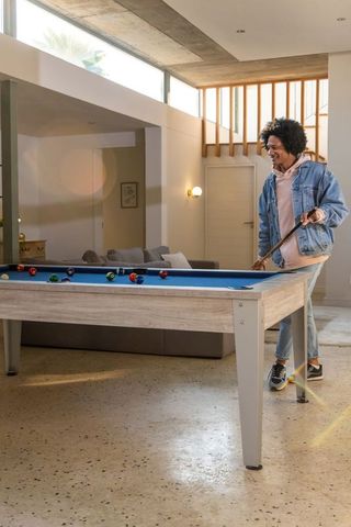 Casual young african american man playing pool at home on blue-felt table in modern living room