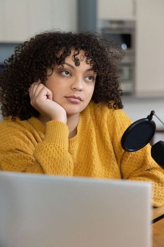 Contemplative woman in mustard sweater recording podcast from home office with laptop mic