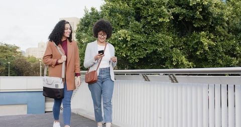 Two friends walking in urban park checking smartphone laughing in casual denim and blazers