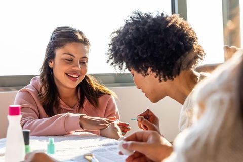 Sunlit diverse friends enjoying at-home manicure painting nails, laughing and bonding