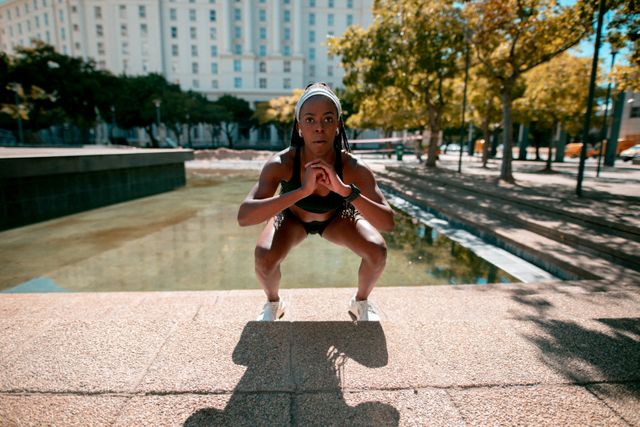African American woman wearing earphones is exercising in a city park. She is performing squats, showcasing her strength and determination. This image is ideal for promoting healthy lifestyles, fitness programs, and outdoor activities. It can be used in advertisements, fitness blogs, and social media campaigns focused on health and wellness.