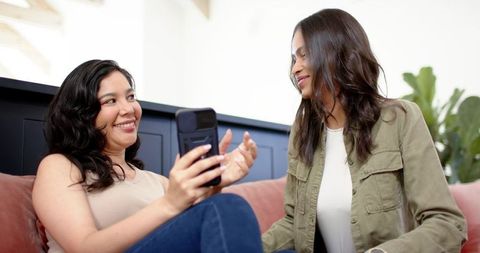 Diverse women sharing smartphone on rose sofa, laughing and chatting in sunlit living room