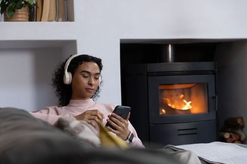 Nonbinary adult relaxing on sofa with headphones and smartphone by fireplace