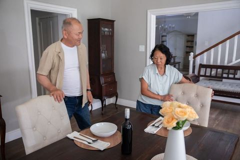 Asian couple arranging tufted dining chairs for cozy home dinner with wine and roses