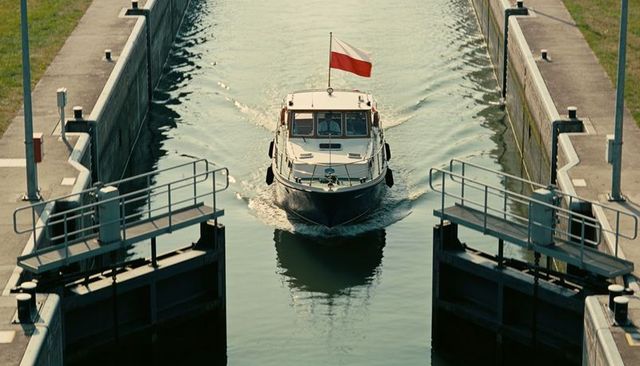 Motorboat navigating canal lock with polish flag, creating v-shaped wake and reflections