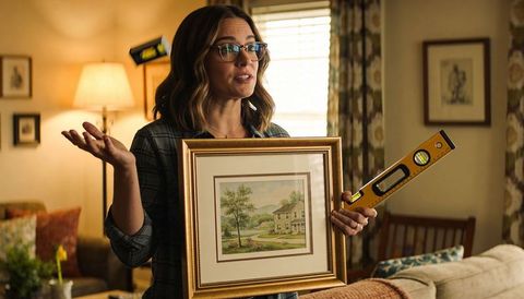 Woman presenting framed landscape while holding yellow spirit level in cozy living room