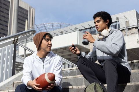 Diverse young men sitting on urban stairs in hoodies holding football and water bottle