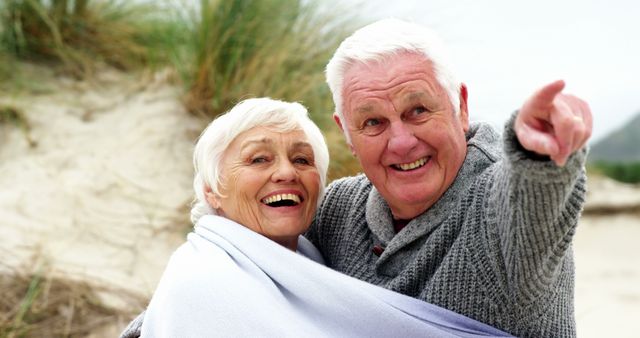 A senior Caucasian couple shares a joyful moment on a beach, wrapped in a blanket, with the man pointing at something in the distance, with copy space. Their smiles and closeness suggest a deep bond and a lifetime of shared memories.