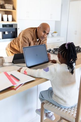 Indian father leaning over kitchen island mentoring african american daughter using laptop