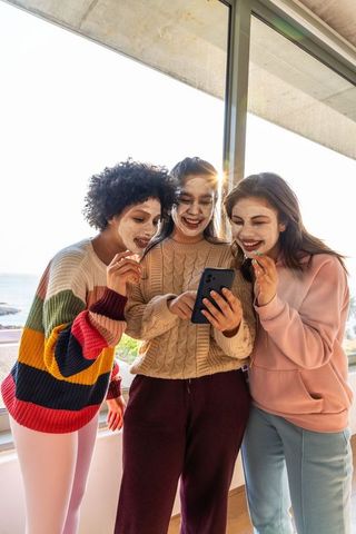 Diverse friends laughing and using smartphone while wearing facial masks near balcony