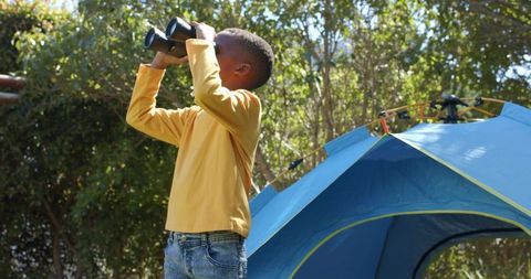 Boy exploring outdoor with binoculars next to blue pop-up tent camping adventure in nature