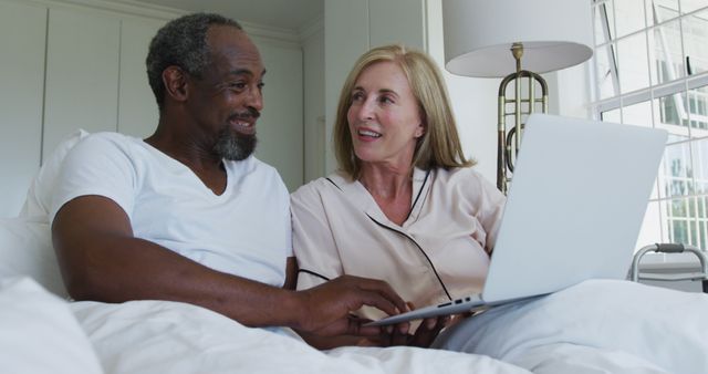 African American man and Caucasian woman are using laptop on bed, engaging each other with smiles. Perfect for illustrating technology use among seniors, promoting digital inclusion, showing relaxed moments, or advertising bed or bedroom products.
