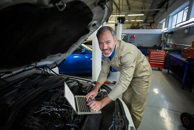 Mechanic in uniform using laptop for diagnostics while servicing car engine in repair shop. Ideal for content related to automotive repair, technology in mechanics, professional services, and car maintenance tutorials.